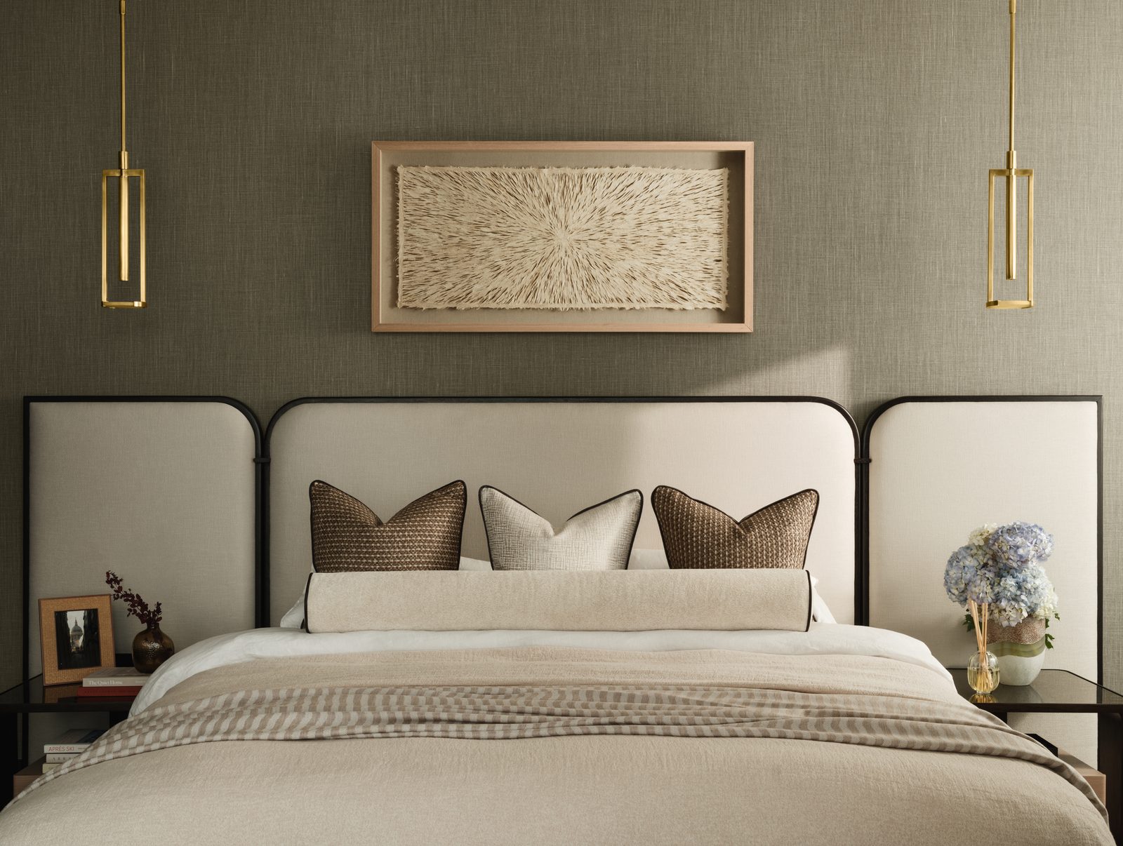 A staged bedroom with an upholstered headboard, layered bedding and a framed textile artwork above the bed
